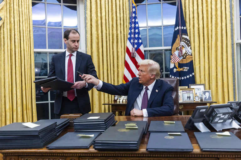 Donald Trump behind the Resolute Desk, hands a pen and some folders to an aide.