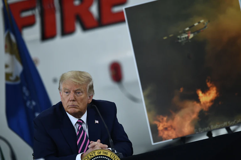 Trump sits at a meeting about wildfires, with an image of a helicopter dumping water on a wildfire in the background.