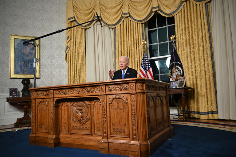 Joe Biden is seen seated at the Resolute Desk in the Oval Office; a boom mike is visible above.