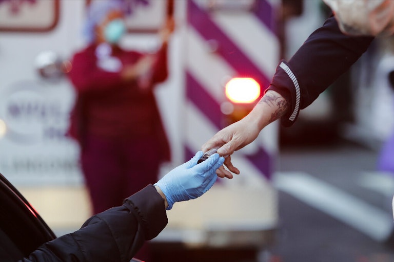 Person in a car handing bottle of hand sanitizer to a person wearing a nitrile glove, an ambulance and health care provider blurred in the background