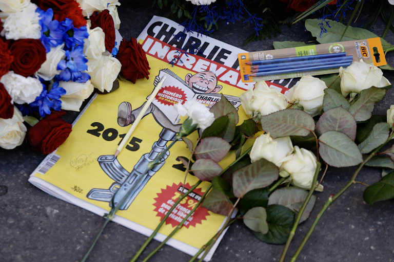 A magazine featuring a cartoon figure seated on a gun, laughing while reading, lies amidst wreaths and pencils left in front of it.