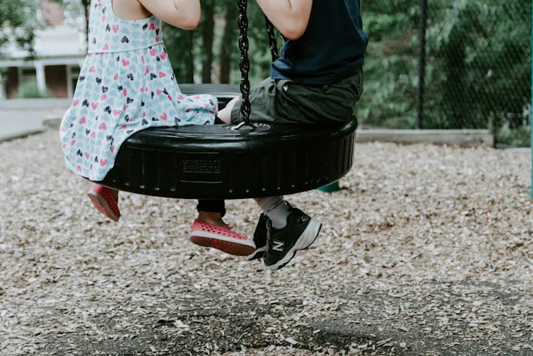 Two children sit on a tyre swing outside.