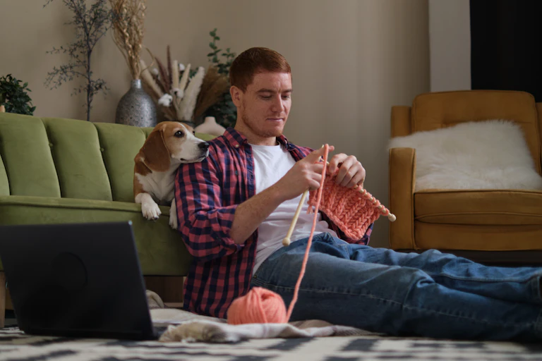 A man knits while sitting on the floor. His beagle sits on the couch behind him.