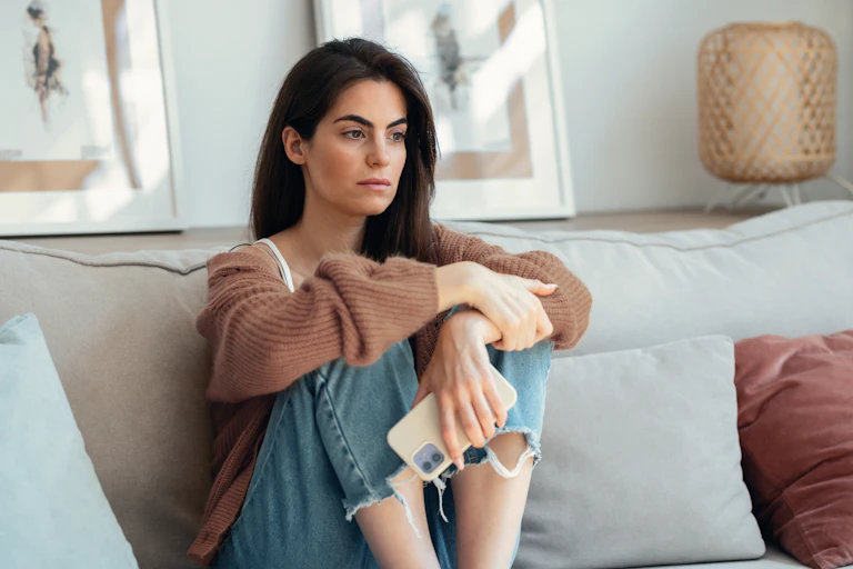 a woman perches on a beige sofa, holding a smartphone and staring into the distance