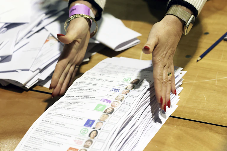 A woman's hands with long red nails reaches for a pile of ballots.
