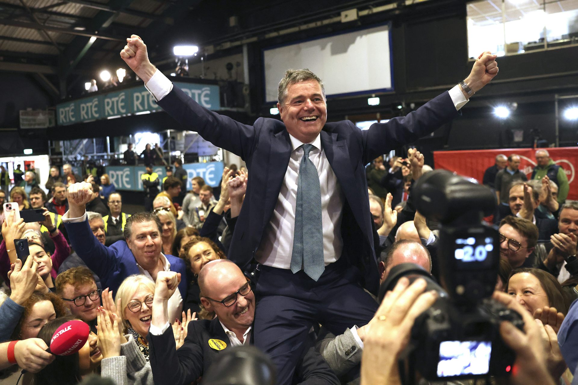 A smiling man with his arms raised on the shoulders of smiling supporters.