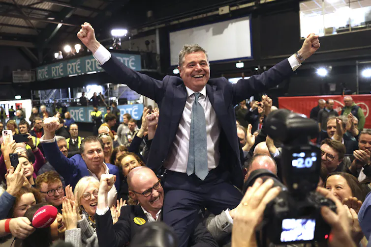 A smiling man with his arms raised on the shoulders of smiling supporters.