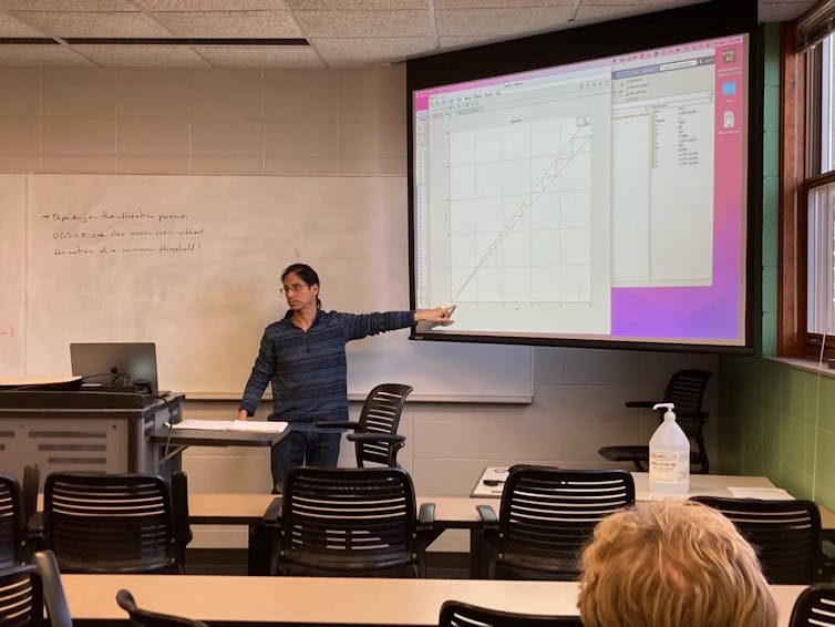 A professor standing in the front of a classroom, pointing at a screen projector.