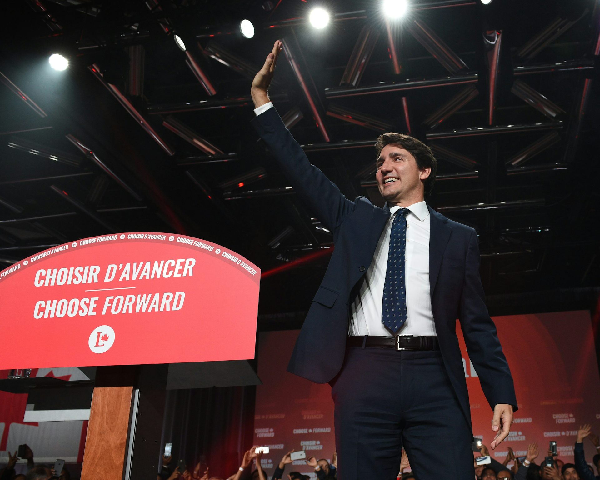 A dark-haired man in a suit smiles and waves next to a red banner that reads Choose Forward in both French and English.