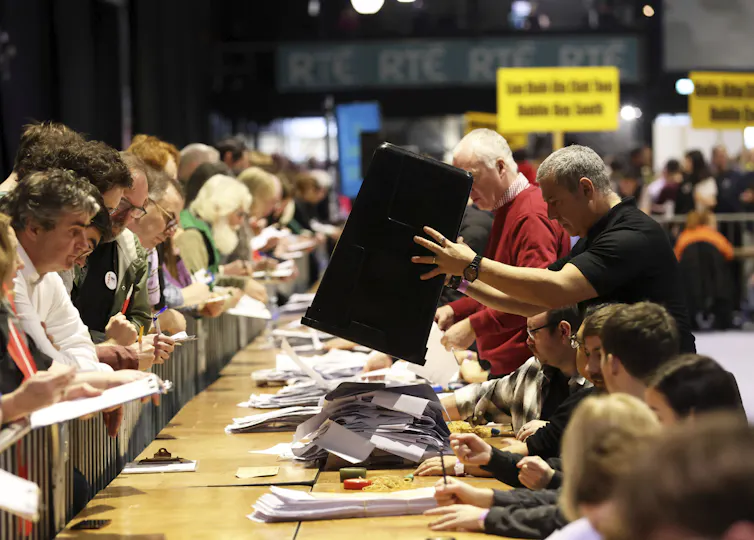 A man dumps a bin of ballots on a long table were ballot counters sit.