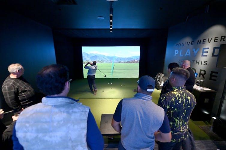 A group of men observe another man participating in a golf simulator.