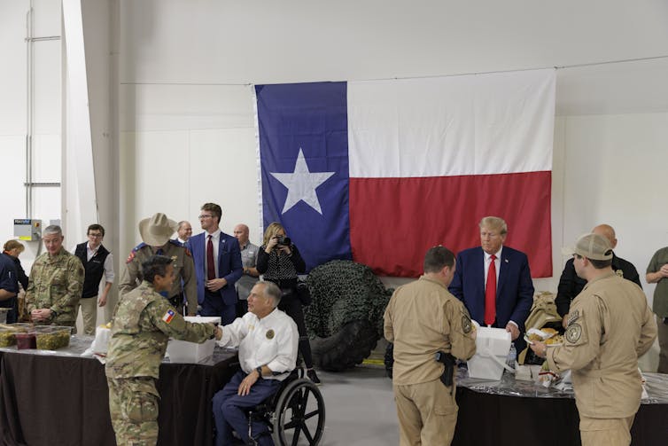 Abbot, in a wheelchair, and President Donald Trump, standing, greet soldiers in uniform in what appears to be an airport hanger