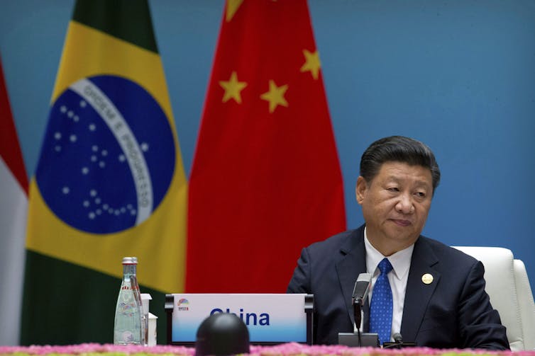 An Asian man sits at a table with flags behind him.