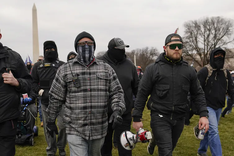 Two men walk in front of a group of masked men with the Washington Monument in the background.