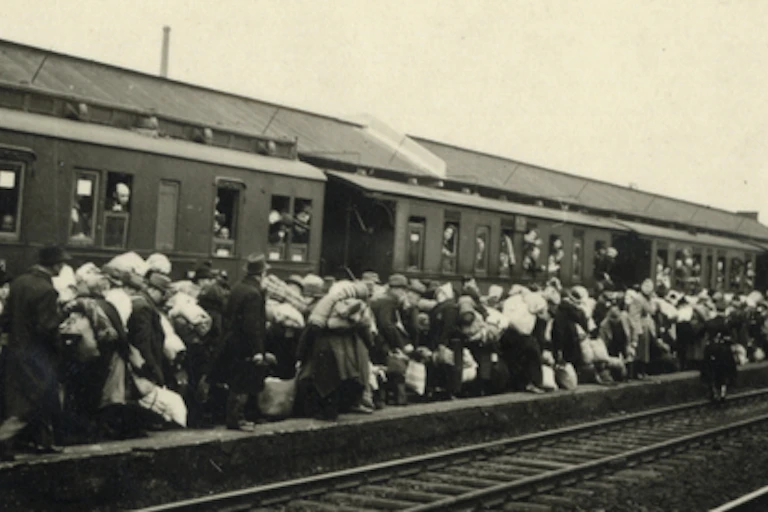 Black-and-white image of people carrying bundles of their belongings on a train platform
