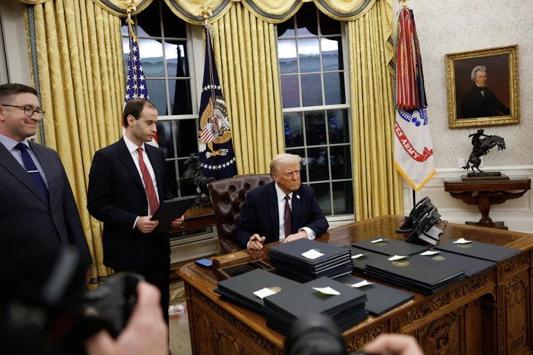 A man holding a pen at a big desk with flags behind him and many blue folders on the desk.