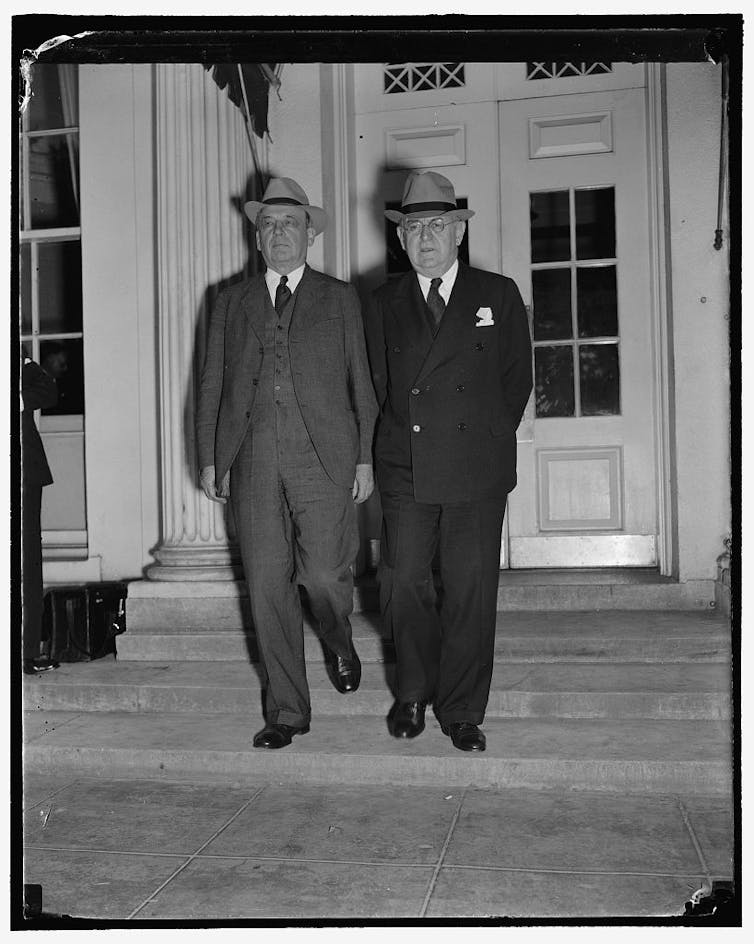 Two men in dark suits and fedoras leave a formal-looking building.