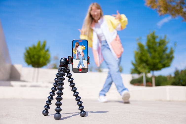 A smartphone on a mount on the ground shows an image of a young woman doing a dance move on a sunny day outside.
