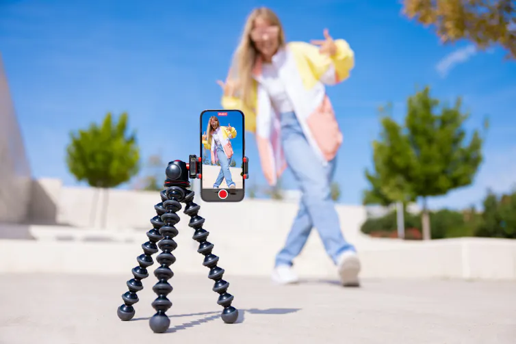 A smartphone on a mount on the ground shows an image of a young woman doing a dance move on a sunny day outside.