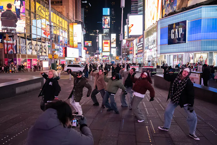 A group of people are seen moving on a busy New York City street, with many bright lights and billboards around them on the buildings.