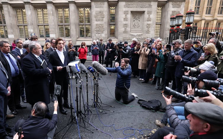 A large media crowd gathers in a semicircle around men speaking into microphones