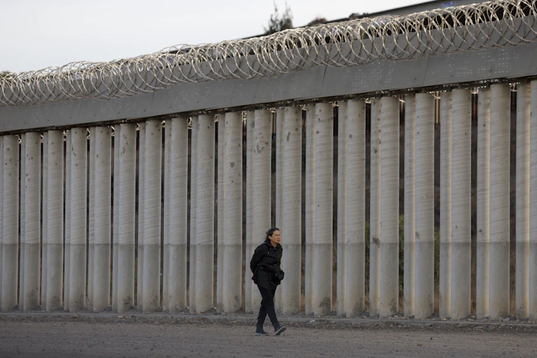 A woman walks along a high wall toppped with razor wire.