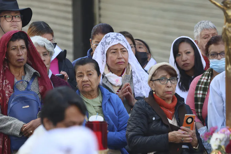 People standing on a street, mostly women, appear to watch something passing by.