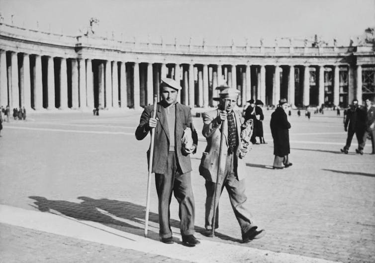 A black and white photograph shows two men with hats and walking sticks crossing a large plaza.