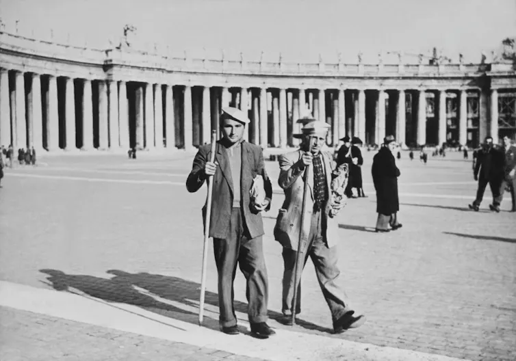 A black and white photograph shows two men with hats and walking sticks crossing a large plaza.