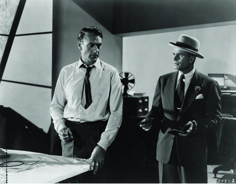 Two men speak in front of drawings, in what appears to be an architect's studio.