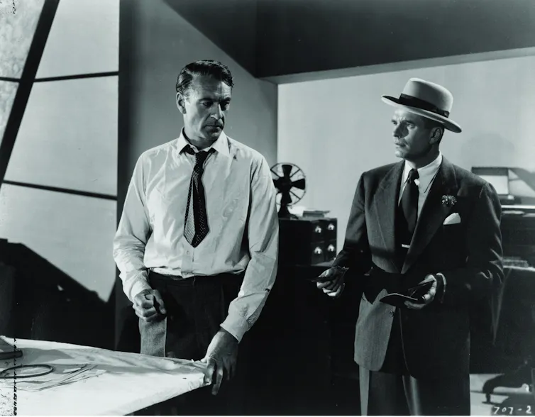 Two men speak in front of drawings, in what appears to be an architect's studio.
