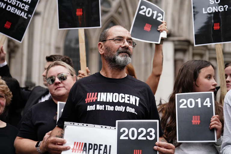 Andrew Malkinson surrounded by supporters in front of the royal courts of justice, wearing a shirt that says 'innocent and not the only one'