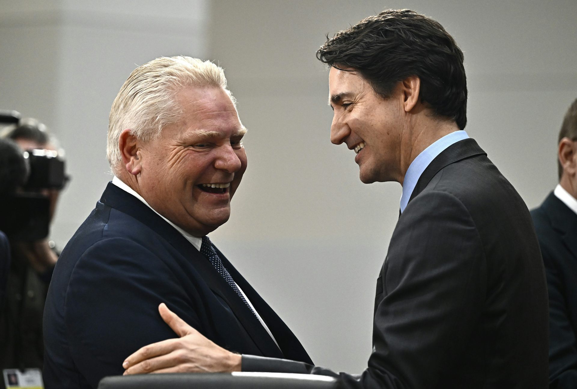 A dark-haired man smiles as he greets a shorter, more rotund man.
