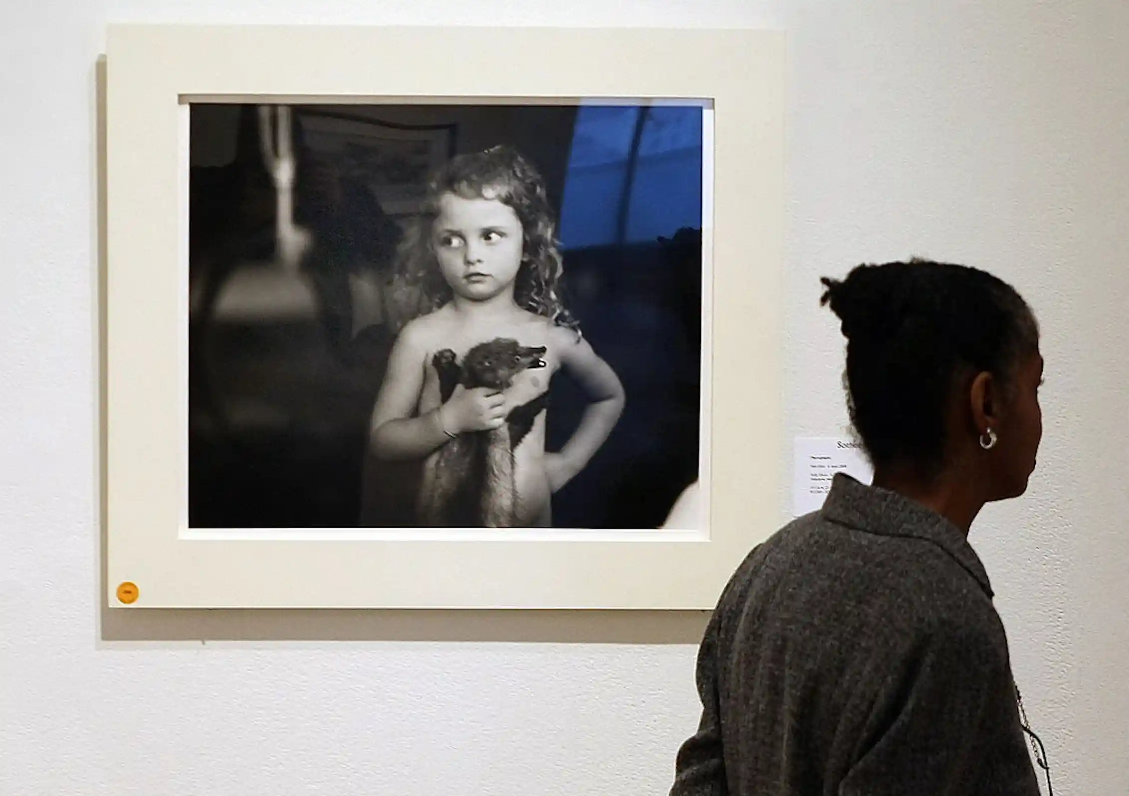 Woman walks by a black and white photograph on display of a young girl holding an weasel by the neck.