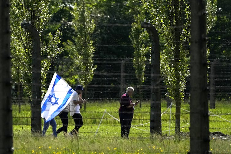 People walk through a grassy forest clearing behind a wire fence. One carries an Israeli flag.