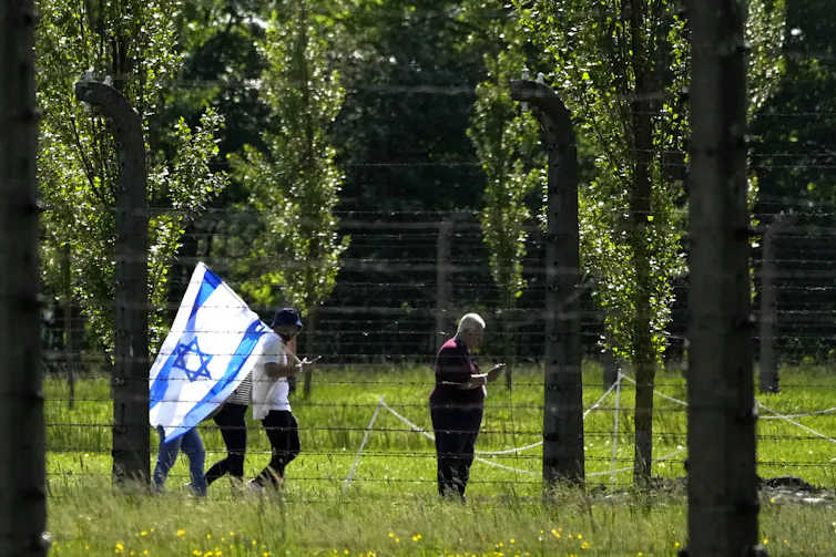 People walk through a grassy forest clearing behind a wire fence. One carries an Israeli flag.