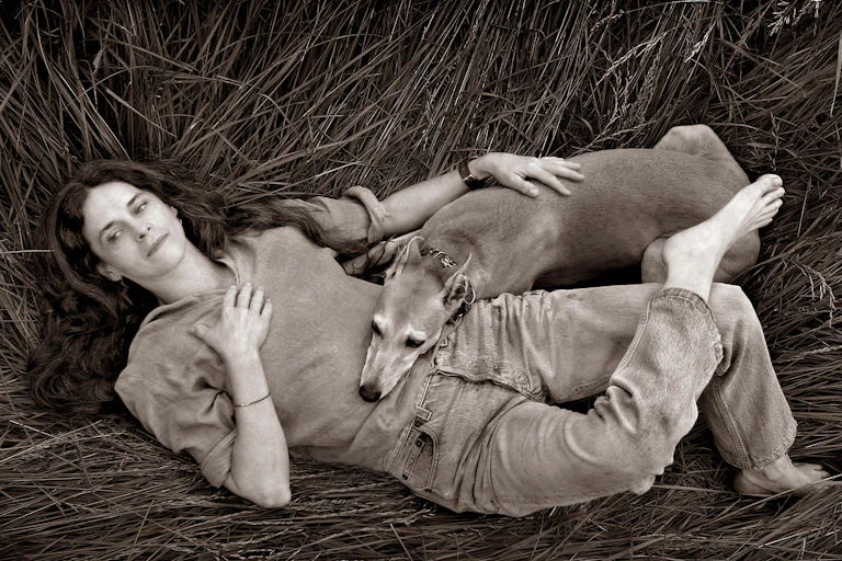 Woman lies in wheat field with a dog snuggled up against her.