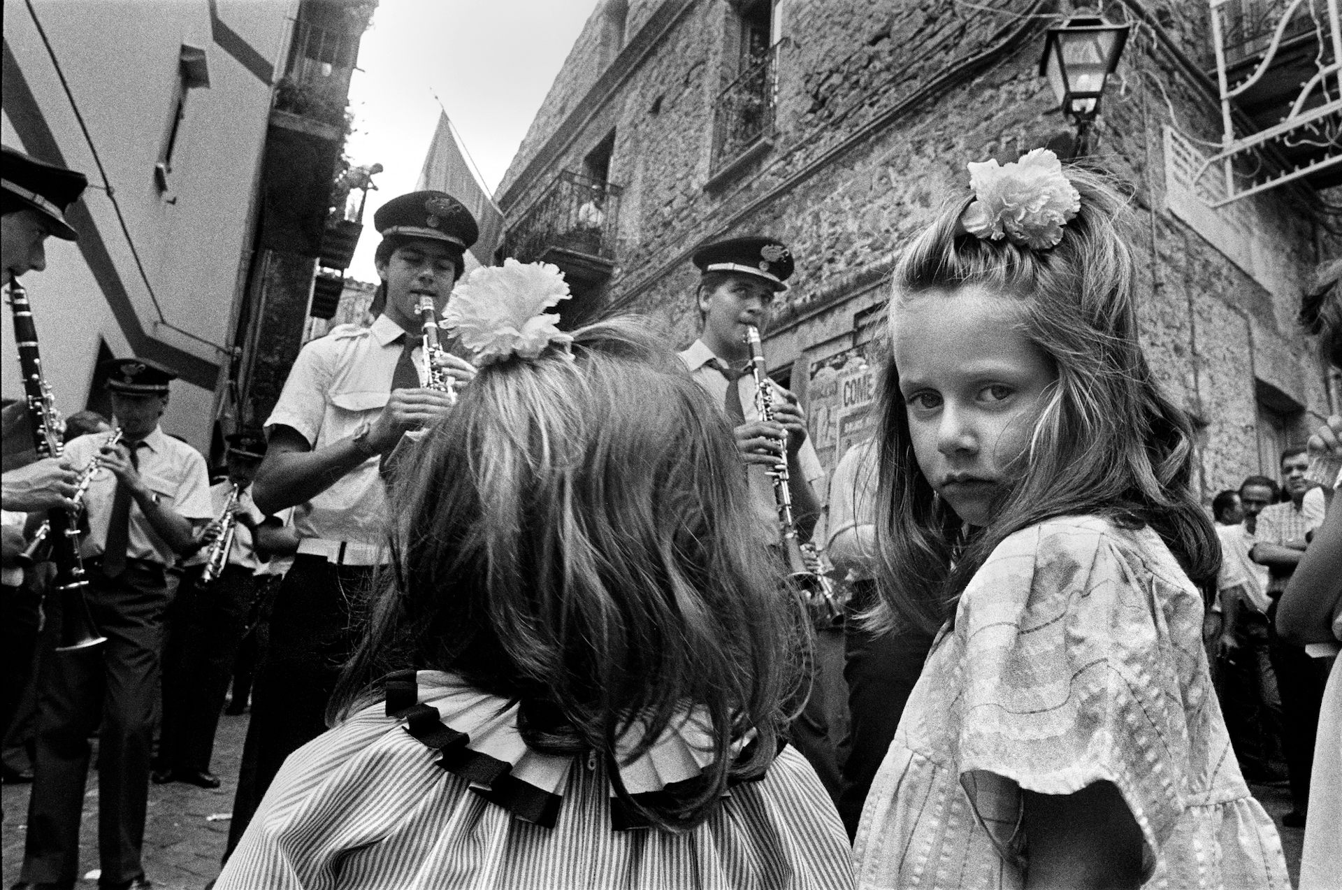 Black and white photograph of two young girls watching an orchestra perform on the street. One of the girls has her head turned to look at the camera.