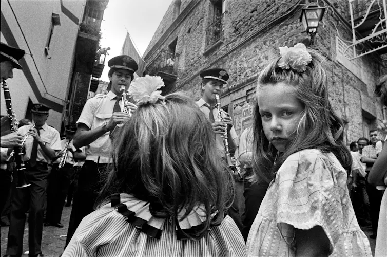 Black and white photograph of two young girls watching an orchestra perform on the street. One of the girls has her head turned to look at the camera.