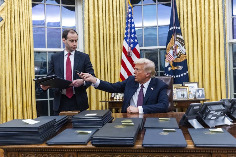 Donald Trump behind the Resolute Desk, hands a pen and some folders to an aide.