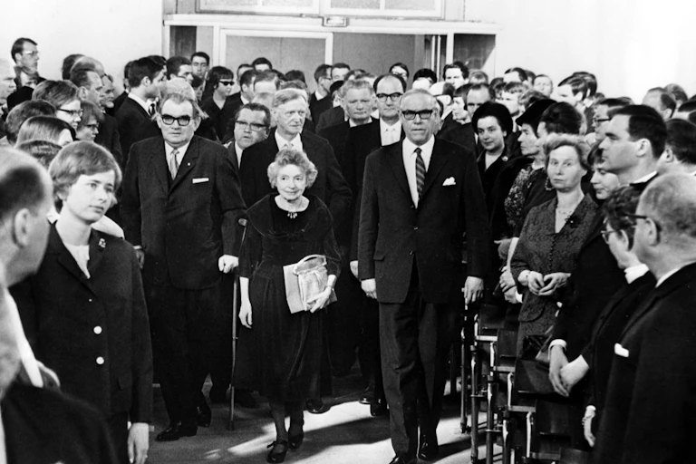 A room of people standing as a shorter woman, Nelly Sachs, walks between them.