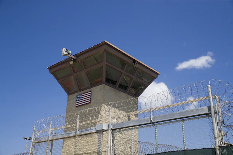 A large tower with windows and a camera stands behind a tall fence topped with barbed wire.