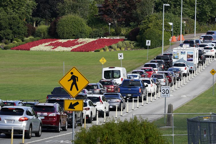 A long line of cars heading in one direction, with a green space and a Canadian flag made of flowers in the background
