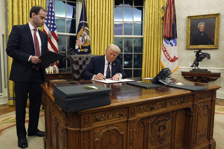 A man sitting at an ornate desk signing a document while another man in a dark suit and red tie stands beside him