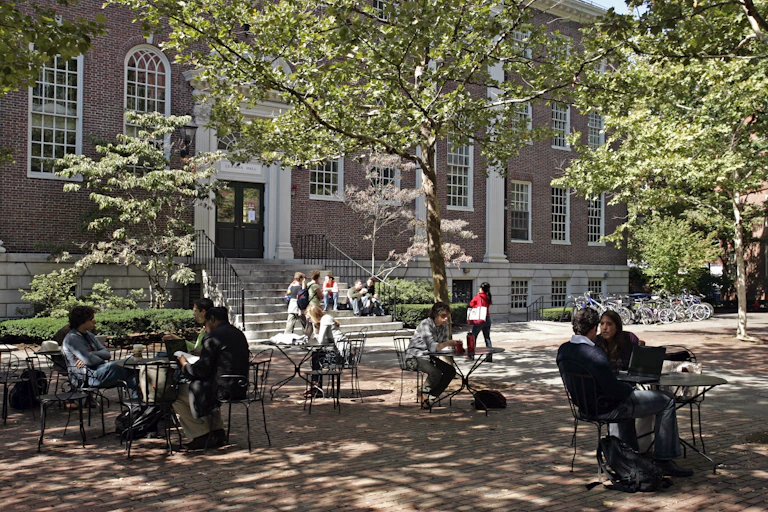 Several students are seated on chairs in the space outside a red brick building.