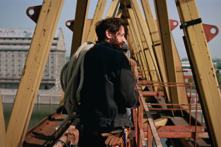 A bearded man on a construction site, high up over a river with a coil of rope over his shoulder.