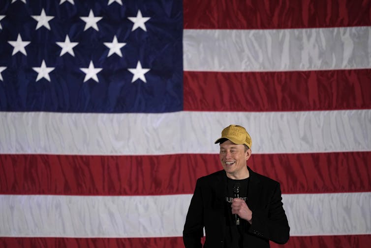 A man in a yellow cap stands in front of an American flag backdrop.