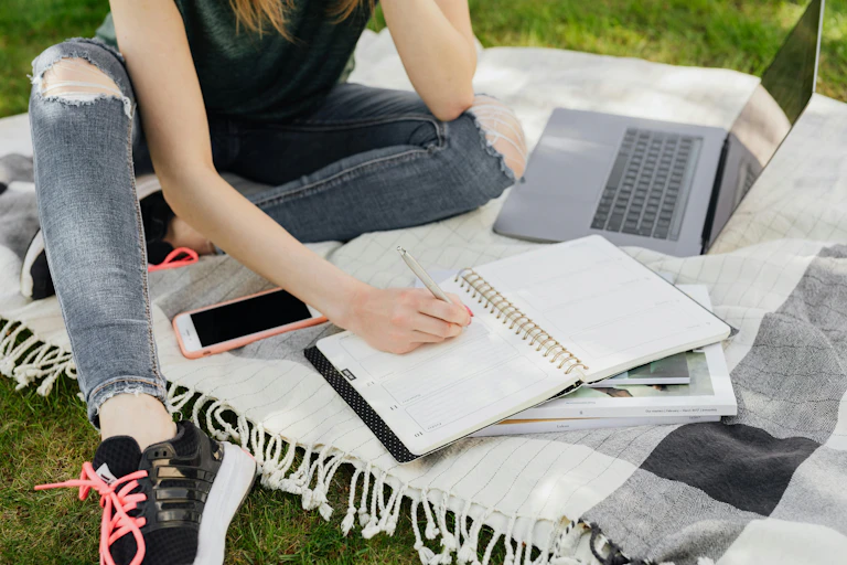 A young person sits on a picnic blanket on grass, with a laptop, planner and notebooks.