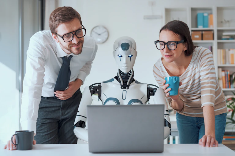 dans un bureau, un robot devant un ordinateur portable entouré d'un homme et d'une femme qui regardent l'écran
