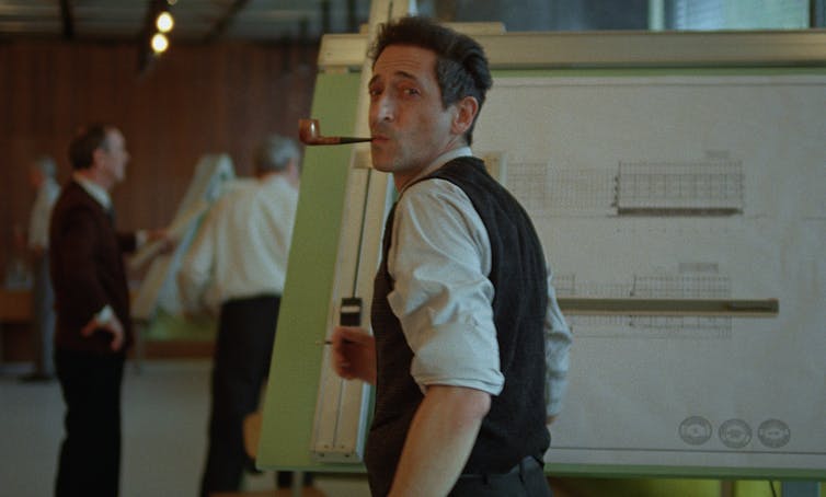 The Brutalist: an architect’s tackle a movie about one guy’s adventure to grasp his visionary construction 3 A man at a drawing board with a pipe in his mouth, looking happy.